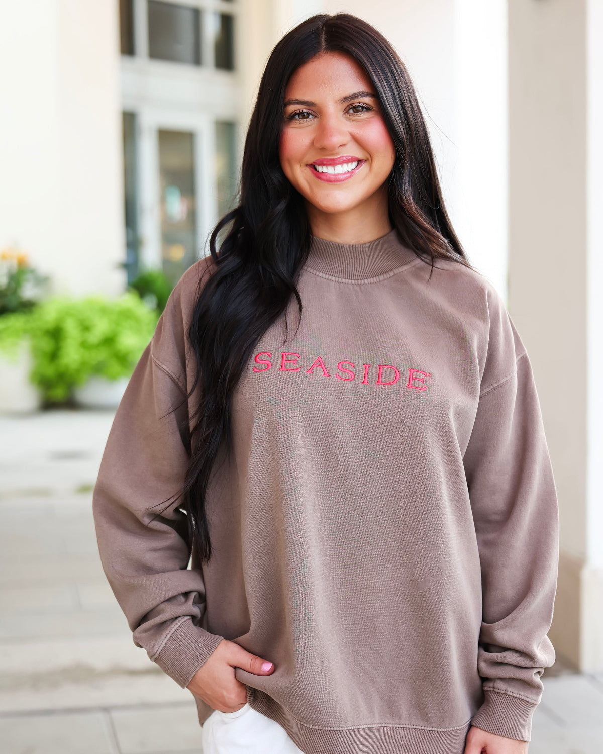 A smiling woman with long dark hair wearing an oversized taupe sweatshirt with 'SEASIDE' embroidered in pink across the chest, standing outdoors in front of a light-colored building.