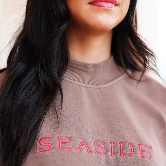 Close-up of a woman wearing a taupe-brown crewneck sweatshirt with 'SEASIDE' embroidered in bright pink across the chest.