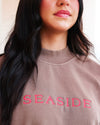 Close-up of a woman wearing a taupe-brown crewneck sweatshirt with 'SEASIDE' embroidered in bright pink across the chest.