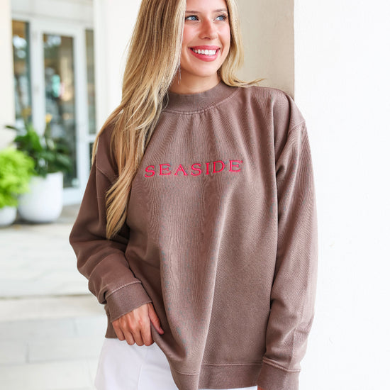 A woman wearing a brown crewneck sweatshirt with 'SEASIDE' embroidered in pink across the chest, paired with white shorts, standing in a bright, modern indoor setting.