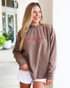 A woman wearing a brown crewneck sweatshirt with 'SEASIDE' embroidered in pink across the chest, paired with white shorts, standing in a bright, modern indoor setting.