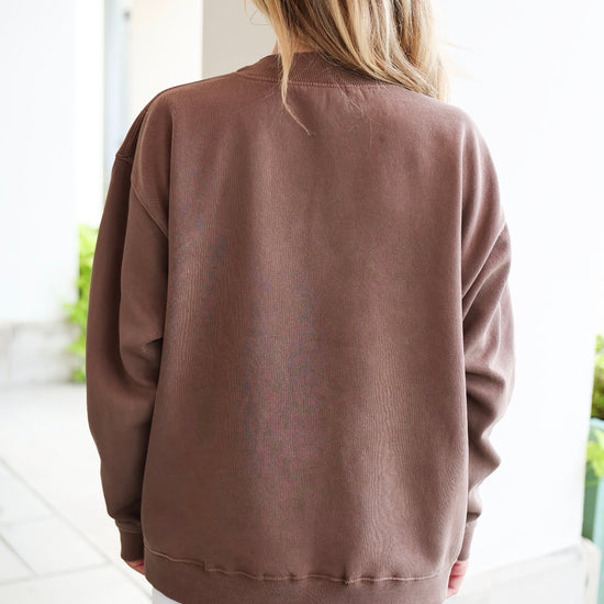 A woman viewed from behind wearing an oversized brown crewneck sweatshirt and white shorts in a bright, modern indoor setting.