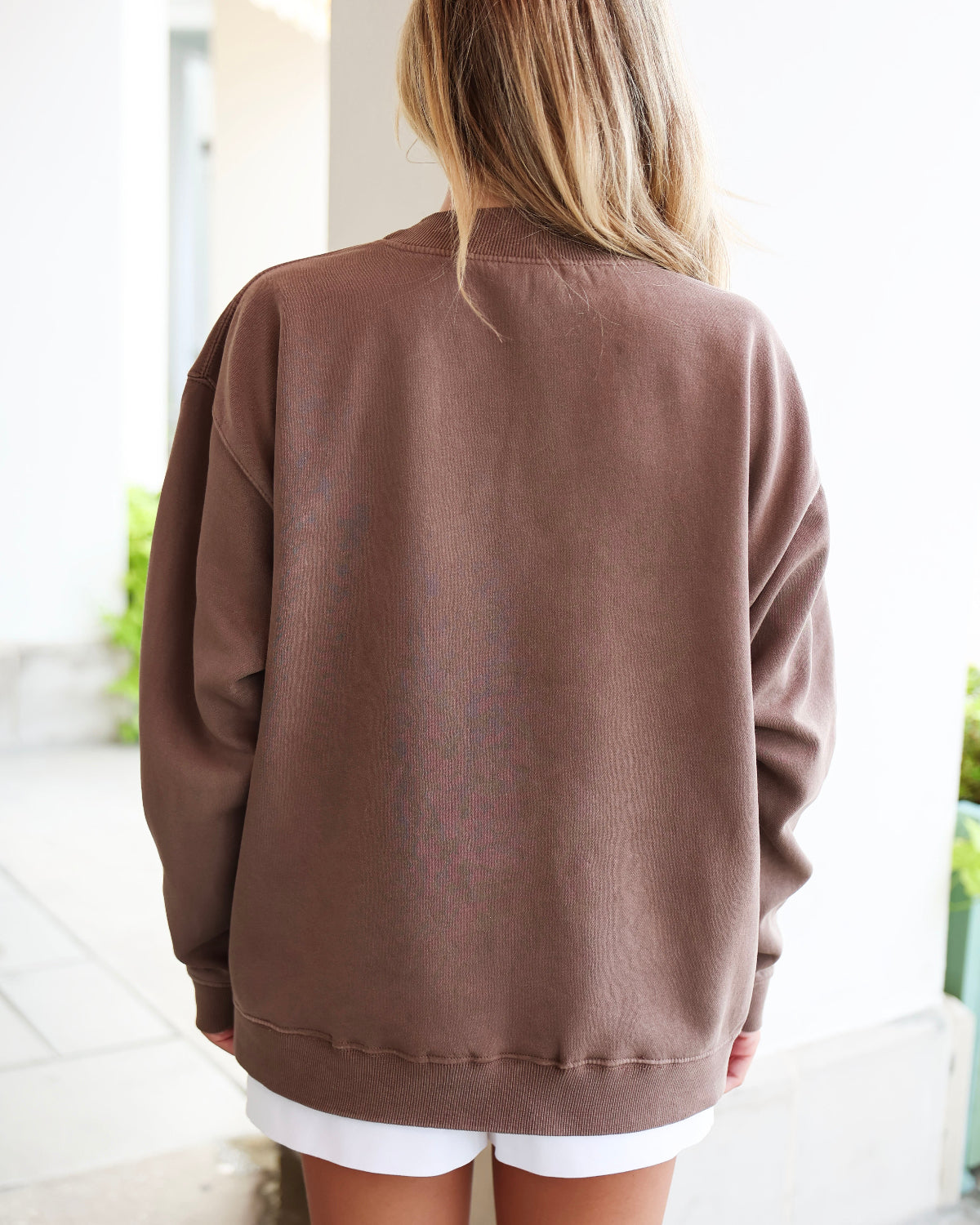 A woman viewed from behind wearing an oversized brown crewneck sweatshirt and white shorts in a bright, modern indoor setting.