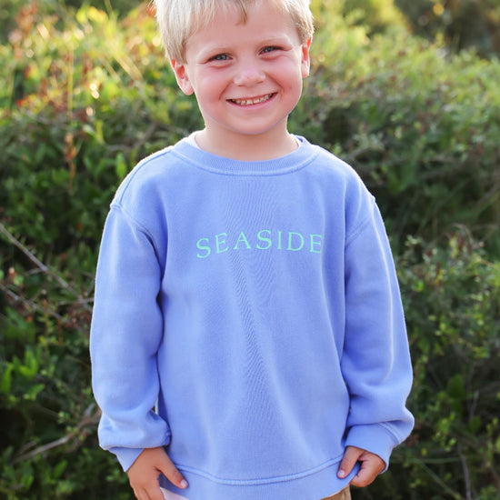 A young blonde boy smiling outdoors, wearing a florida blue crewneck sweatshirt with 'SEASIDE' printed in light green across the chest, paired with tan pants.