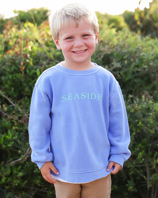A young blonde boy smiling outdoors, wearing a florida blue crewneck sweatshirt with 'SEASIDE' printed in light green across the chest, paired with tan pants.