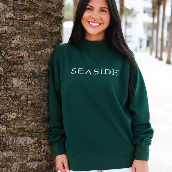 A woman wearing a dark green mock neck sweatshirt with 'SEASIDE' printed in white across the chest, standing outdoors next to a palm tree trunk.