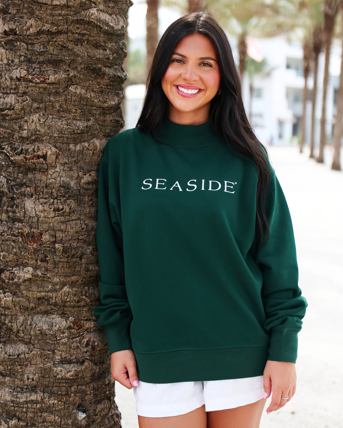 A woman wearing a dark green mock neck sweatshirt with 'SEASIDE' printed in white across the chest, standing outdoors next to a palm tree trunk.