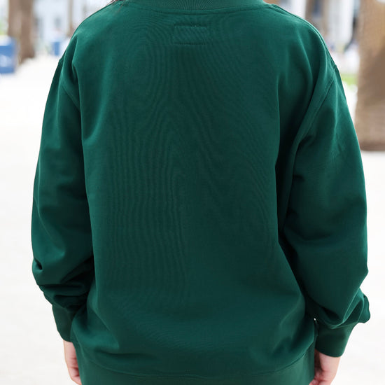 A person viewed from behind wearing a dark forest green long-sleeve sweatshirt in an outdoor setting with blurred trees in the background.