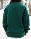 A person viewed from behind wearing a dark forest green long-sleeve sweatshirt in an outdoor setting with blurred trees in the background.