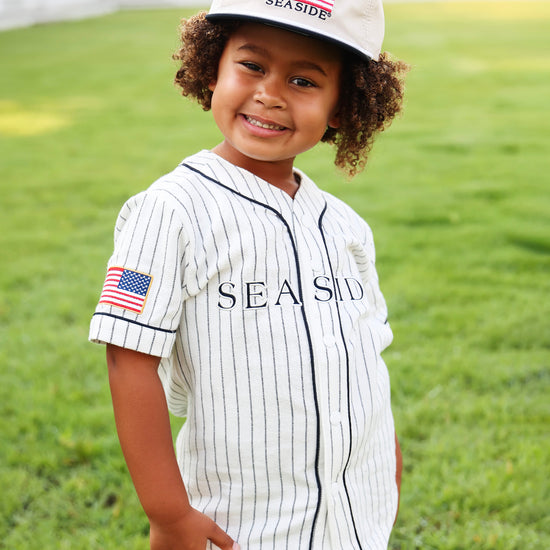 A young child wearing a white pinstripe baseball-style shirt dress with 'SEASIDE' printed across the chest, matching cap with an American flag patch and 'SEASIDE' text, standing on a green grassy lawn.