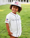 A young child wearing a white pinstripe baseball-style shirt dress with 'SEASIDE' printed across the chest, matching cap with an American flag patch and 'SEASIDE' text, standing on a green grassy lawn.