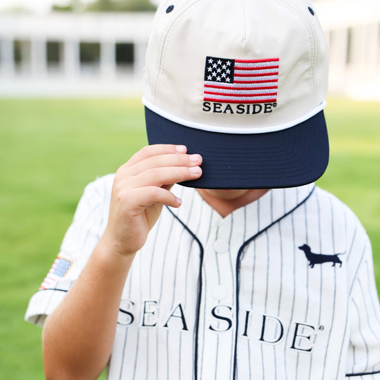 A child wearing a white and navy baseball cap with an embroidered American flag and the word 'SEASIDE' on the front, paired with a matching white pinstripe baseball jersey on a blurred green field background.