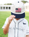 A child wearing a white and navy baseball cap with an embroidered American flag and the word 'SEASIDE' on the front, paired with a matching white pinstripe baseball jersey on a blurred green field background.