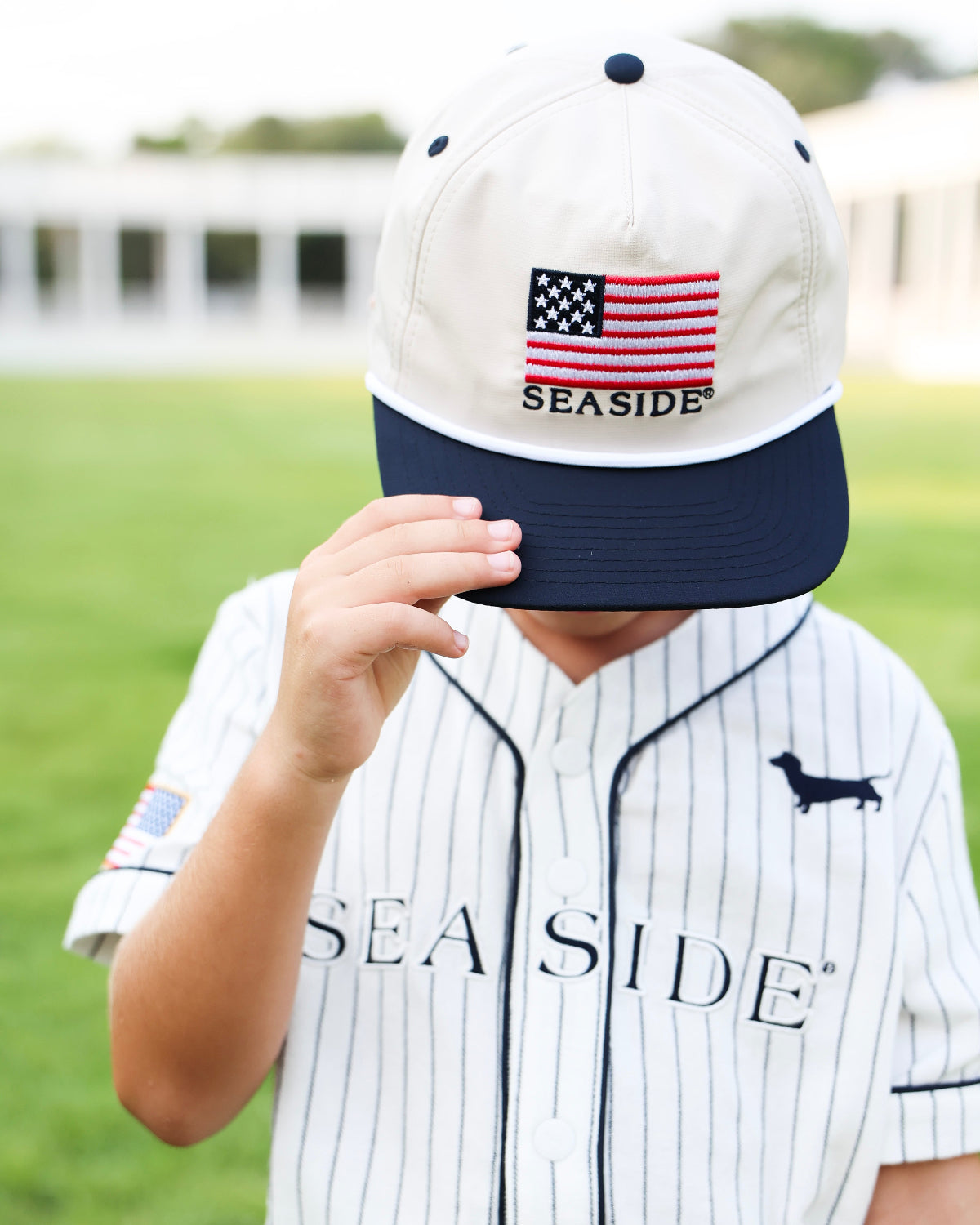 A child wearing a white and navy baseball cap with an embroidered American flag and the word 'SEASIDE' on the front, paired with a matching white pinstripe baseball jersey on a blurred green field background.