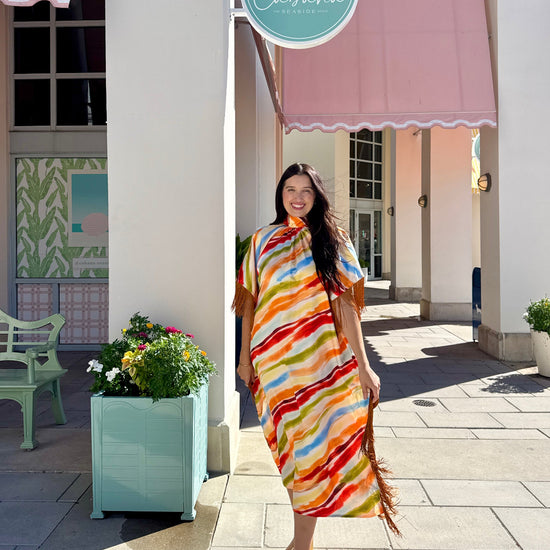 A woman wearing a vibrant, multicolored striped kaftan dress with fringe details walks on a sunny outdoor sidewalk in front of a building with pink awnings and a 'Cabana' sign.