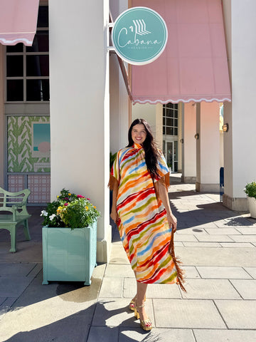 A woman wearing a vibrant, multicolored striped kaftan dress with fringe details walks on a sunny outdoor sidewalk in front of a building with pink awnings and a 'Cabana' sign.