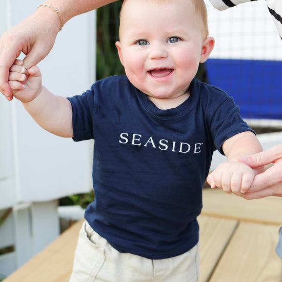 A baby wearing a navy blue t-shirt with 'SEASIDE' printed in white across the chest, walking outdoors while being held up by an adult.