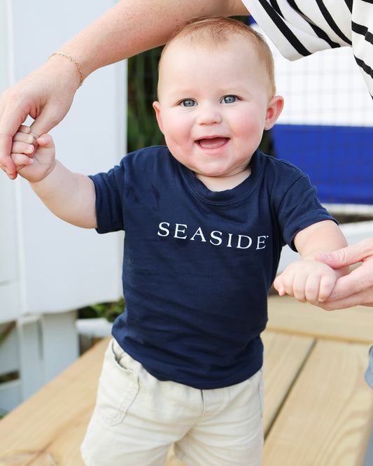 A baby wearing a navy blue t-shirt with 'SEASIDE' printed in white across the chest, walking outdoors while being held up by an adult.