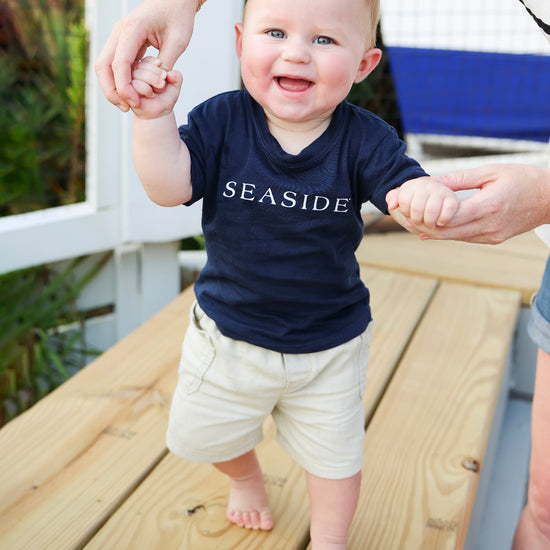 A baby walking barefoot on a wooden deck, wearing a navy blue t-shirt with 'SEASIDE' printed in white across the chest and beige shorts. An adult is holding the baby's hands to assist with balance.