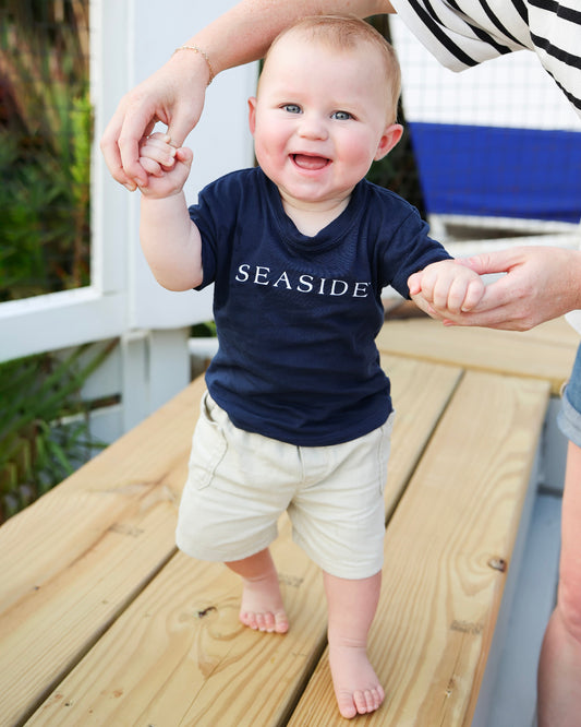 A baby walking barefoot on a wooden deck, wearing a navy blue t-shirt with 'SEASIDE' printed in white across the chest and beige shorts. An adult is holding the baby's hands to assist with balance.