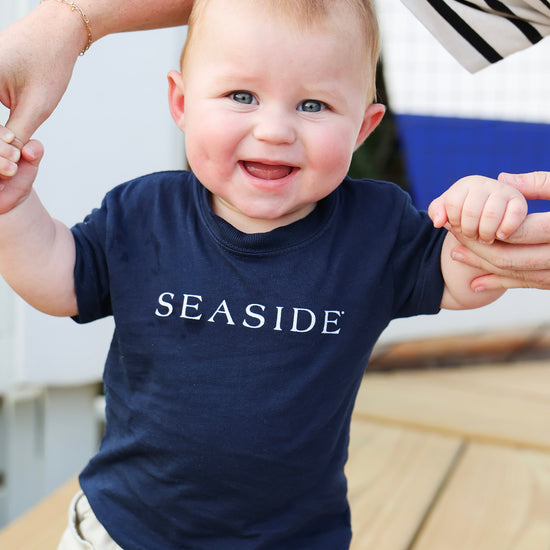 A baby wearing a navy blue t-shirt with 'SEASIDE' printed in white across the chest, held up by an adult's hands on a wooden deck.