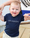 A baby wearing a navy blue t-shirt with 'SEASIDE' printed in white across the chest, held up by an adult's hands on a wooden deck.