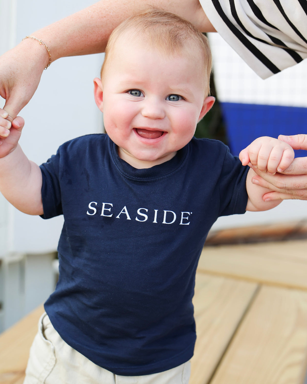A baby wearing a navy blue t-shirt with 'SEASIDE' printed in white across the chest, held up by an adult's hands on a wooden deck.
