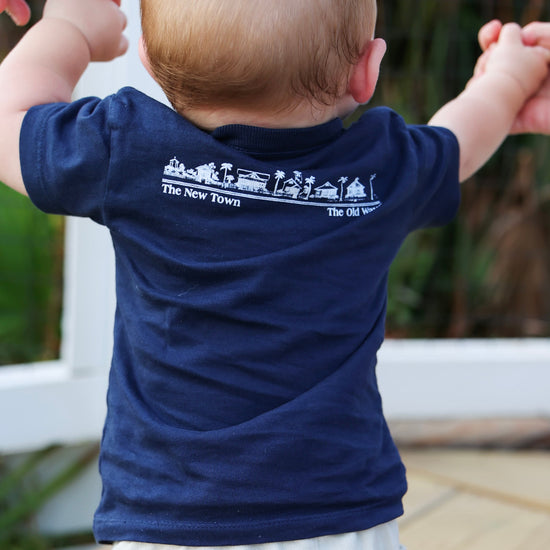A baby viewed from behind wearing a navy blue short-sleeve t-shirt with a white graphic print on the back that reads 'The New Town' and 'The Old Ways'.