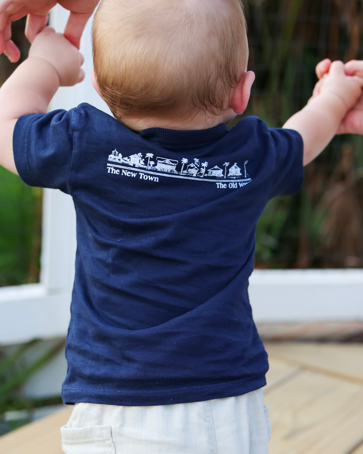 A baby viewed from behind wearing a navy blue short-sleeve t-shirt with a white graphic print on the back that reads 'The New Town' and 'The Old Ways'.