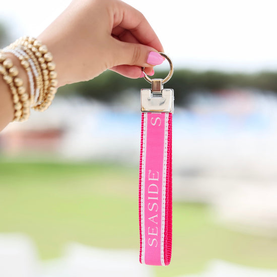 A hand with pink nail polish and gold bracelets holds a bright pink Seaside Keychain strap with a silver metal ring, set against a blurred outdoor background.