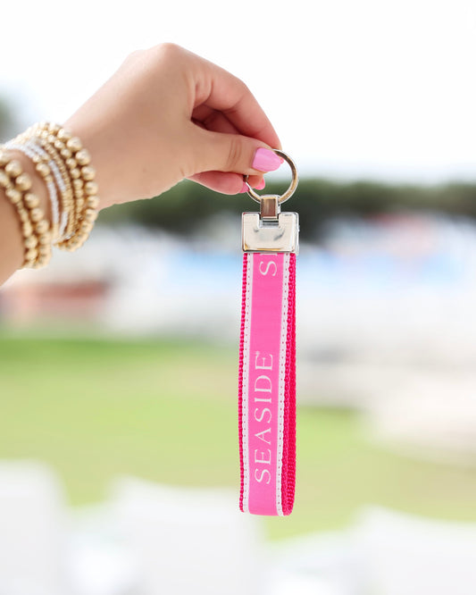 A hand with pink nail polish and gold bracelets holds a bright pink Seaside Keychain strap with a silver metal ring, set against a blurred outdoor background.