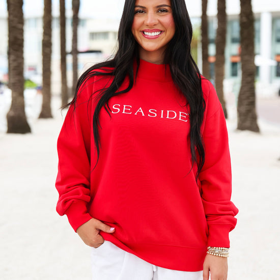 A woman with long black hair stands outdoors wearing a bright red crewneck sweatshirt with 'SEASIDE' printed in white across the chest, paired with white shorts.