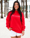 A woman with long black hair stands outdoors wearing a bright red crewneck sweatshirt with 'SEASIDE' printed in white across the chest, paired with white shorts.
