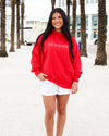 A woman stands on a beach walkway wearing a bright red crewneck sweatshirt with 'SEASIDE' printed in white across the chest, paired with white shorts and clear high-heel sandals.