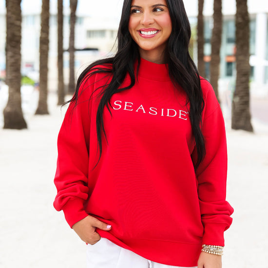 A woman wearing a bright red crewneck sweatshirt with 'SEASIDE' printed in white across the chest, paired with white shorts, standing outdoors on a beach boardwalk with palm trees in the background.