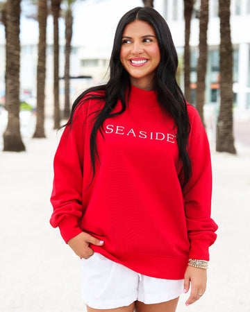 A woman wearing a bright red crewneck sweatshirt with 'SEASIDE' printed in white across the chest, paired with white shorts, standing outdoors on a beach boardwalk with palm trees in the background.