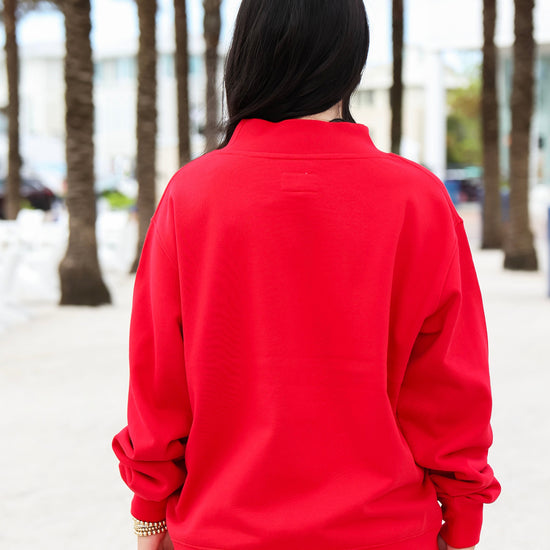A person viewed from behind wearing a bright red crewneck sweatshirt and white pants, standing outdoors on a sandy path lined with palm trees.