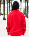 A person viewed from behind wearing a bright red crewneck sweatshirt and white pants, standing outdoors on a sandy path lined with palm trees.
