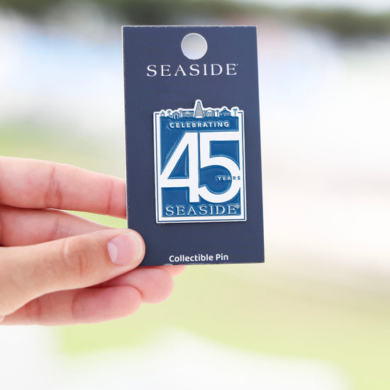 A hand holding a dark blue collectible pin card featuring a Seaside 45th Anniversary pin, with a blurred outdoor background.