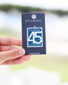 A hand holding a dark blue collectible pin card featuring a Seaside 45th Anniversary pin, with a blurred outdoor background.