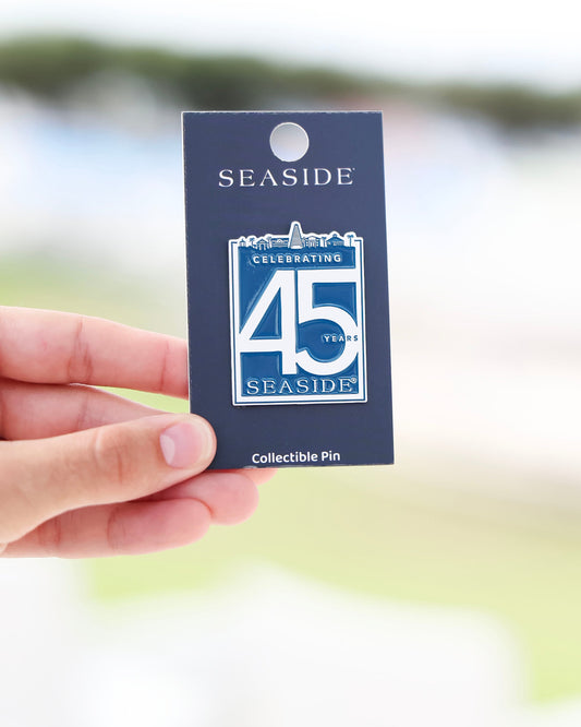A hand holding a dark blue collectible pin card featuring a Seaside 45th Anniversary pin, with a blurred outdoor background.