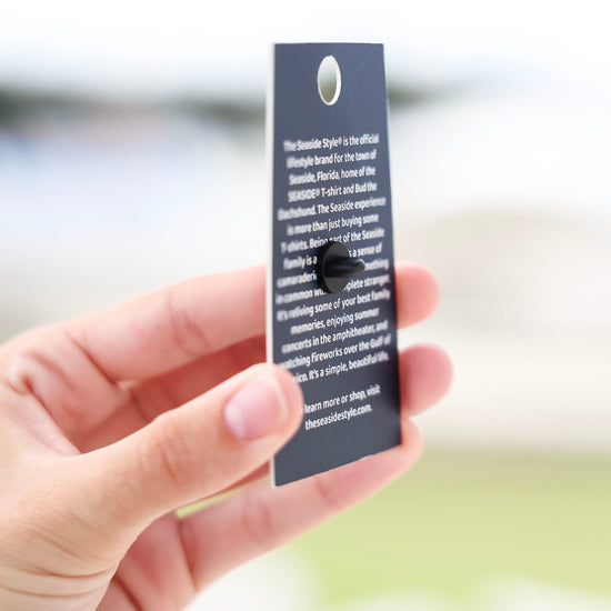 A hand holding a small rectangular printed card or tag with a hole at the top, shown against a softly blurred outdoor background.