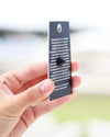 A hand holding a small rectangular printed card or tag with a hole at the top, shown against a softly blurred outdoor background.