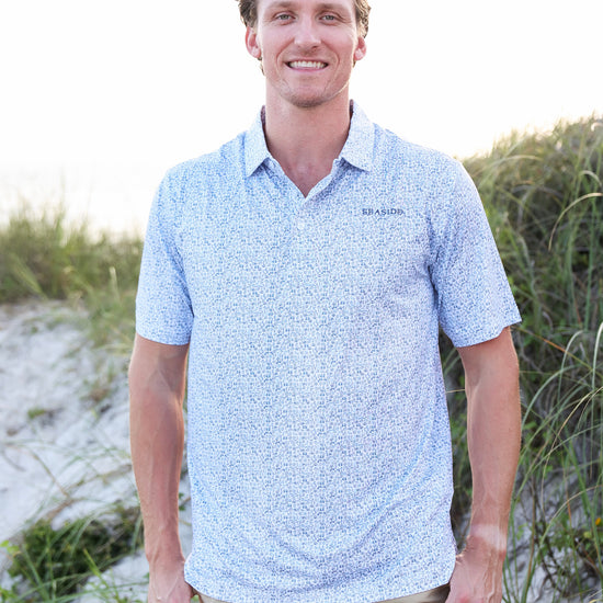 A smiling man wearing a light blue and white micro-patterned short-sleeve polo shirt with khaki pants, standing outdoors on a sandy dune with green grass in the background.
