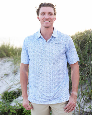 A smiling man wearing a light blue and white micro-patterned short-sleeve polo shirt with khaki pants, standing outdoors on a sandy dune with green grass in the background.