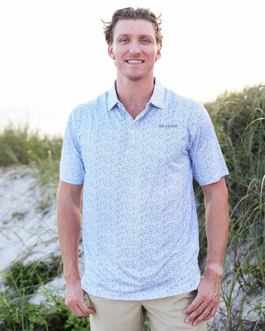 A smiling man wearing a light blue and white micro-patterned short-sleeve polo shirt with khaki pants, standing outdoors on a sandy dune with green grass in the background.