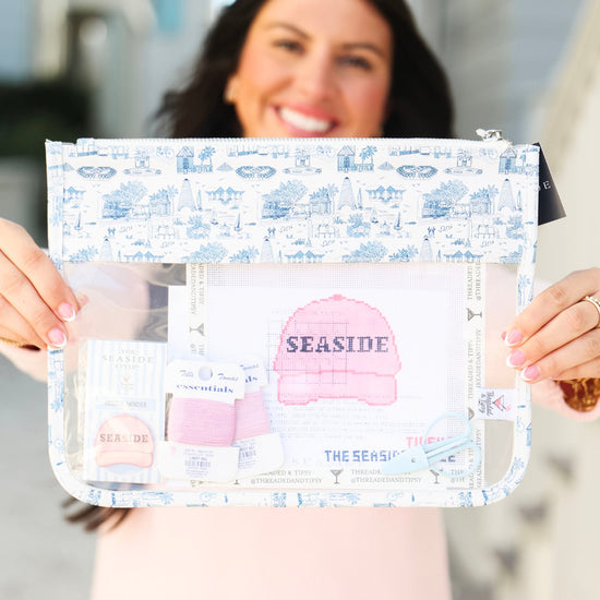 A woman holds up a clear zippered pouch with a blue and white nautical-themed print. The pouch has a transparent front window displaying a pink baseball cap and other small accessories, with the word 'SEASIDE' printed on the cap.