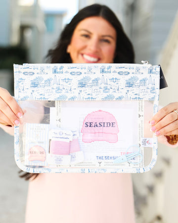 A woman holds up a clear zippered pouch with a blue and white nautical-themed print. The pouch has a transparent front window displaying a pink baseball cap and other small accessories, with the word 'SEASIDE' printed on the cap.