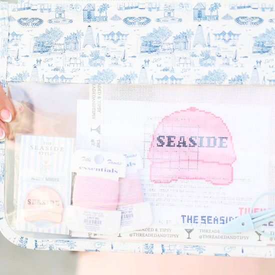 A clear zippered pouch with a blue and white toile de Jouy patterned border, holding various sewing and crafting supplies including a pink Seaside knit hat, thread, and pattern paper. A person is holding the pouch, with their face blurred out of focus in the background.