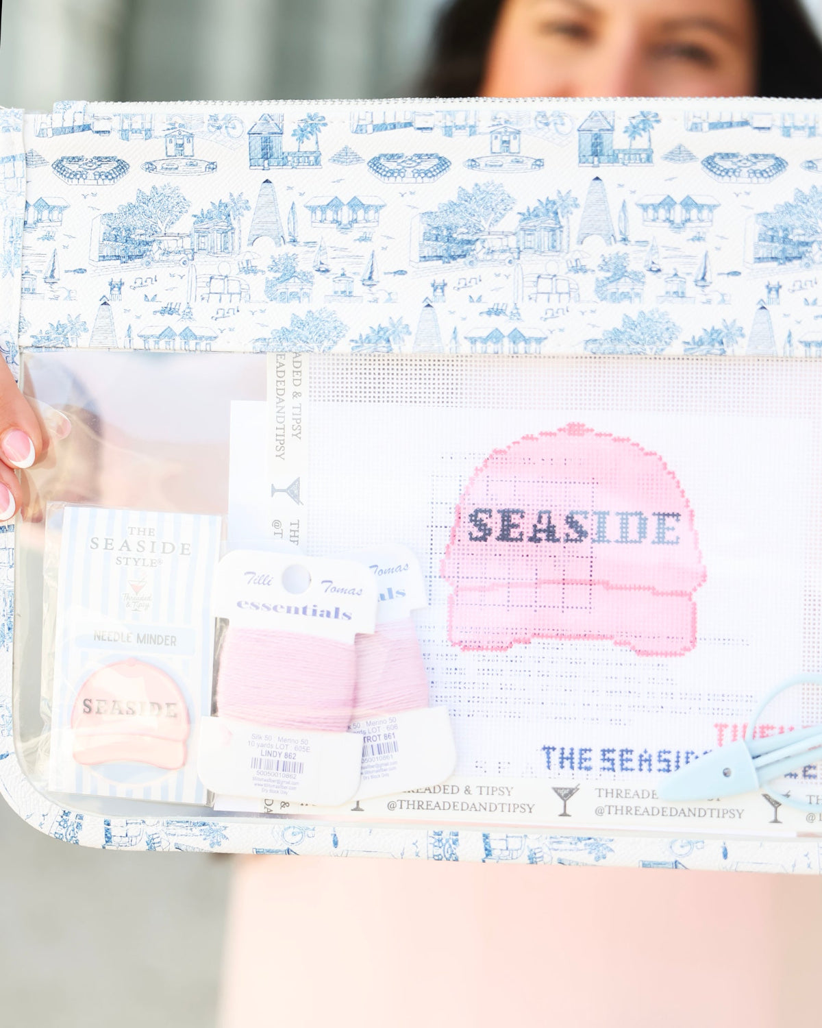 A clear zippered pouch with a blue and white toile de Jouy patterned border, holding various sewing and crafting supplies including a pink Seaside knit hat, thread, and pattern paper. A person is holding the pouch, with their face blurred out of focus in the background.